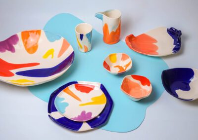 Colorful porcelain plates, bowls, and cups with multicolored brushstroke designs on a light blue background.